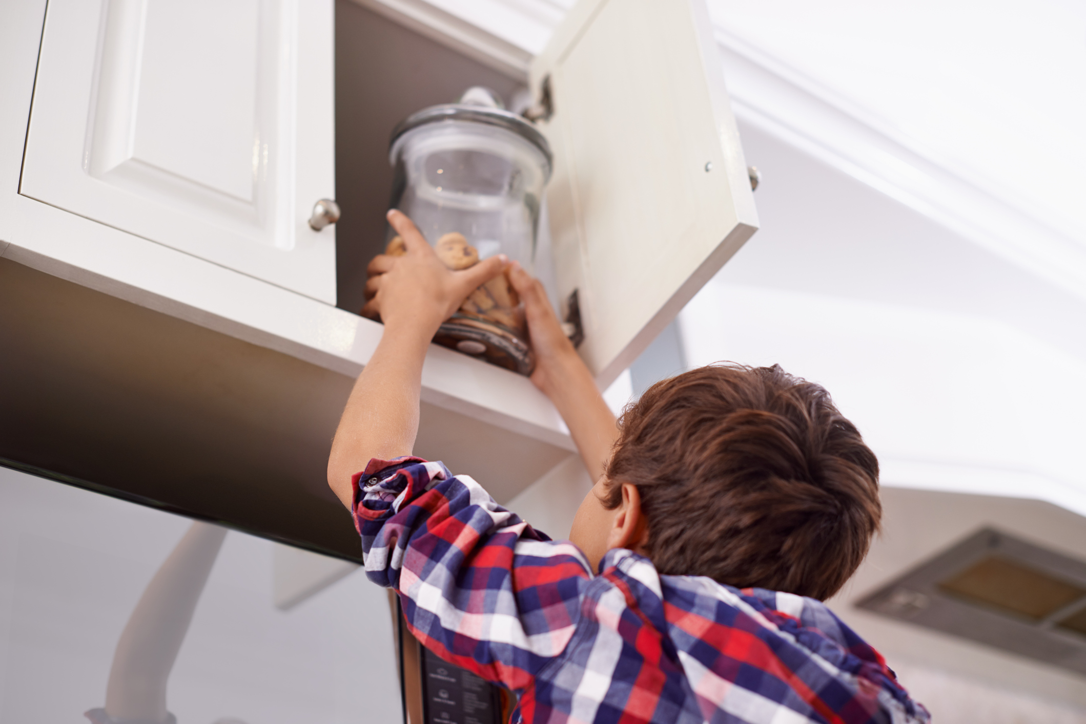 A young boy reaching for a cookie jar in the top kitchen cupboard